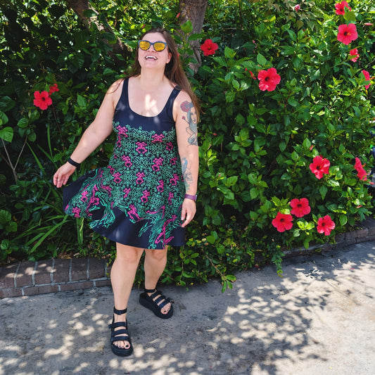 A woman wearing a black sleeveless dress with a green and pink pattern print, standing outdoors in front of a lush green bush with red flowers.