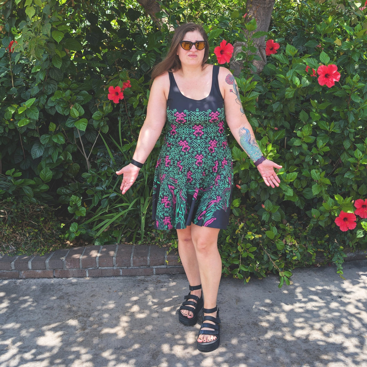 A woman wearing a sleeveless black and green pattern print dress with pink flowers, standing outdoors in front of a lush green garden with red hibiscus flowers.