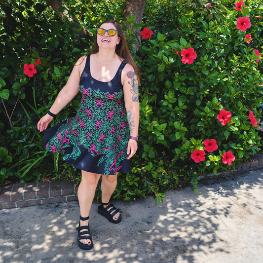 A woman wearing a black sleeveless dress with a green and pink pattern print, standing outdoors in front of a lush green bush with red flowers.